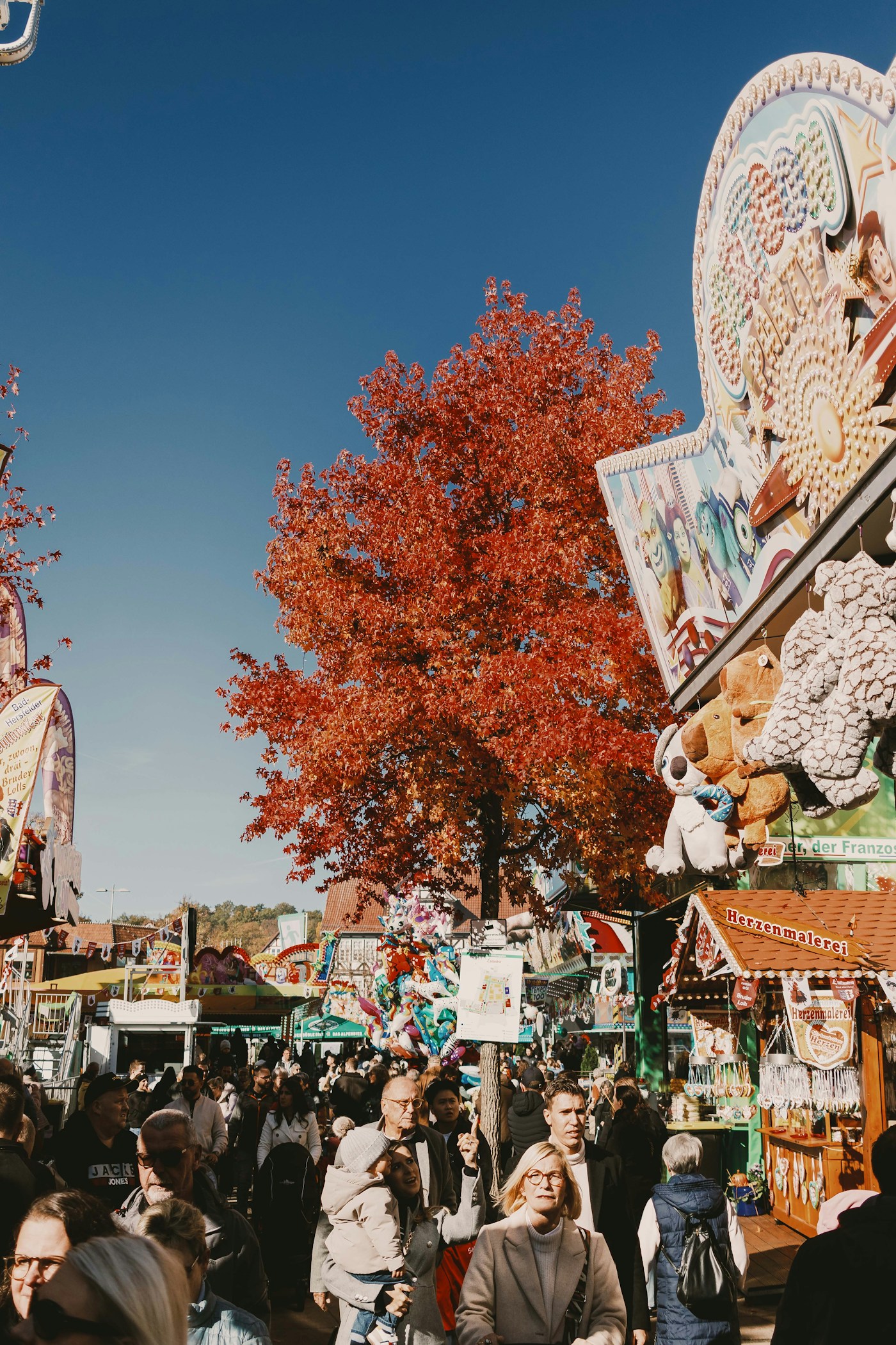 Crowds passing colorful carnival prize stalls beneath an orange autumn tree